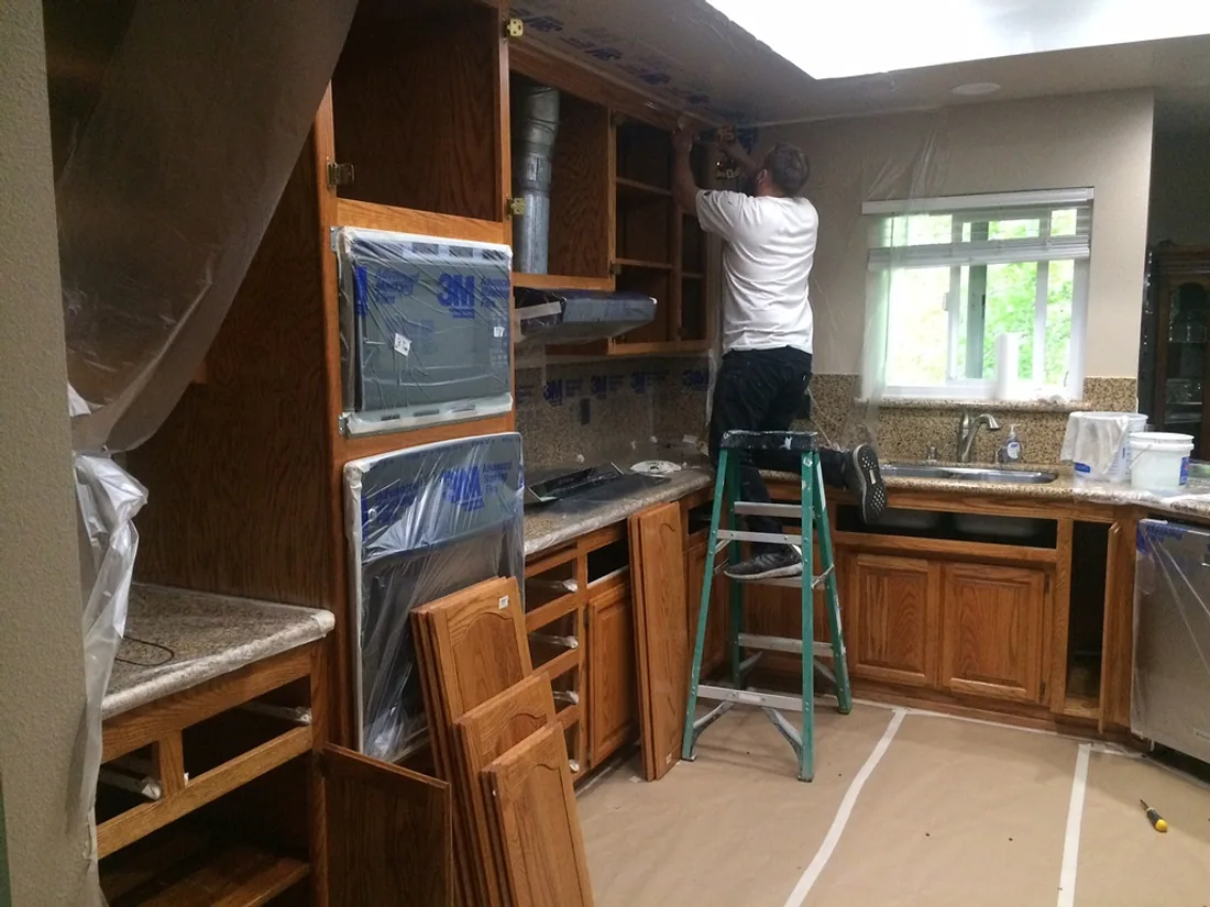 Kitchen cabinets masked and being primed by a painter on a ladder